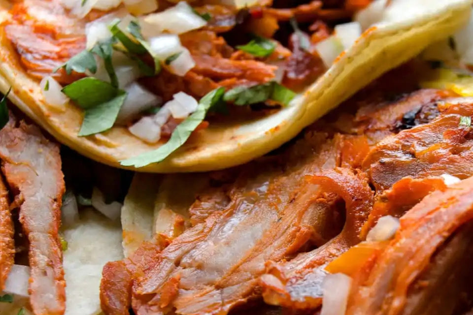 primer plano de detalle de tacos al pastor donde se ve la carne rojiza sobre una tortilla de maíz y aderezado con cebolla y cilantro