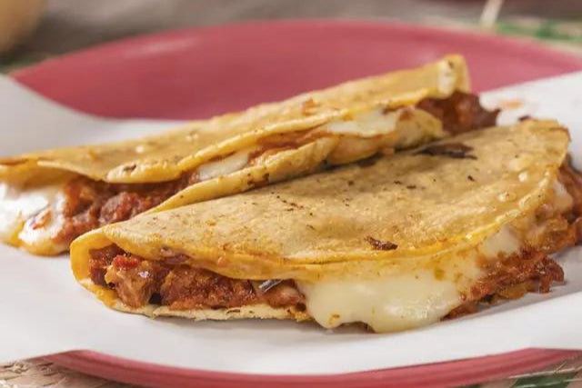 dos quesadillas de las que asoma chicharron rojizo y queso fundidoen un plato rosado sobre una mesa