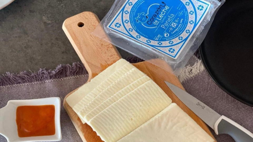 Sobre una encimera gris, tabla de madera con queso cortado en lonchas gruesas, un cuchillo, plato a los lados y el envase del queso panela de la marca corazon de leche