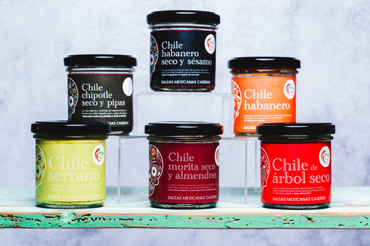 Línea completa de 6 salsas caseras mexicanas en frascos, dispuestas en forma piramidal sobre una tabla azul y fondo blanco
