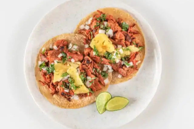 Vista cenital de dos tacos al pastor sobre un plato blanco y con sendos limones verdes cortados