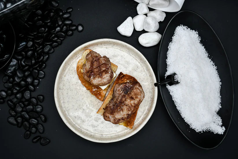 Vista cenital de un plato blanco con montaditos de carne junto a otro ovalado negro con sal blanca, sobre mesa negra y piedras negras y blancas