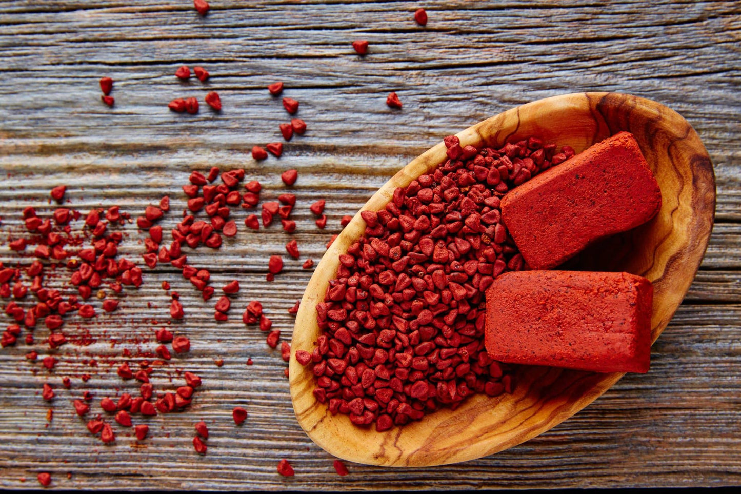 Semillas de achiote esparcidas sobre una mesa de madera, acompañadas de bloques de pasta de achiote en un cuenco rústico.