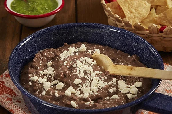 Frijoles negros refritos servidos en un sartén azul, decorados con queso fresco, acompañados de totopos y salsa verde.