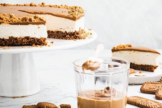 en primer termino un vaso de cafe con leche junto a unas galletas biscoff, detrás una tarta tipo cheesecake cortada y un plato con una porción de tarta.