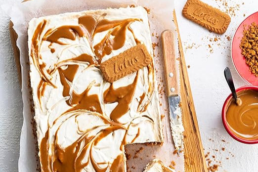 sobre una tabla de madera tarta y migas de galletas biscoff un bol de crema, cuchillo, una galleta y un plato con migas de galleta