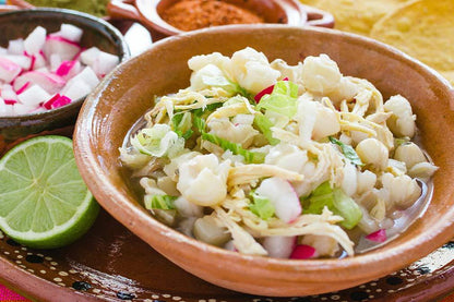 Tazón de pozole con maíz blanco, pollo desmenuzado, lechuga, rábanos, limón y guarniciones.