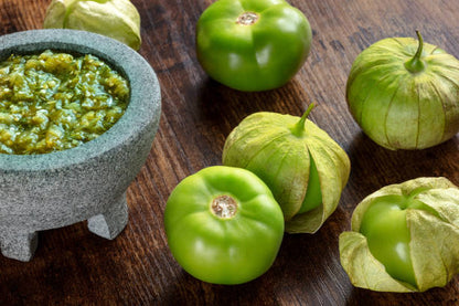 Tomatillos frescos junto a un molcajete con salsa verde casera sobre mesa de madera. Ingrediente clave en la cocina mexicana.