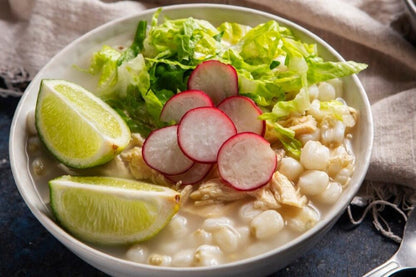 Plato de pozole blanco con maíz, pollo, lechuga, rábanos y rodajas de limón, servido en un tazón blanco.