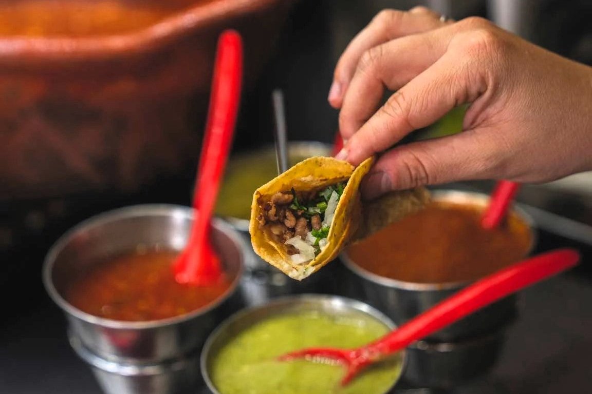 Taco con carne y salsa, tomado con la mano, listo para disfrutar, acompañado de diferentes salsas tradicionales mexicanas.